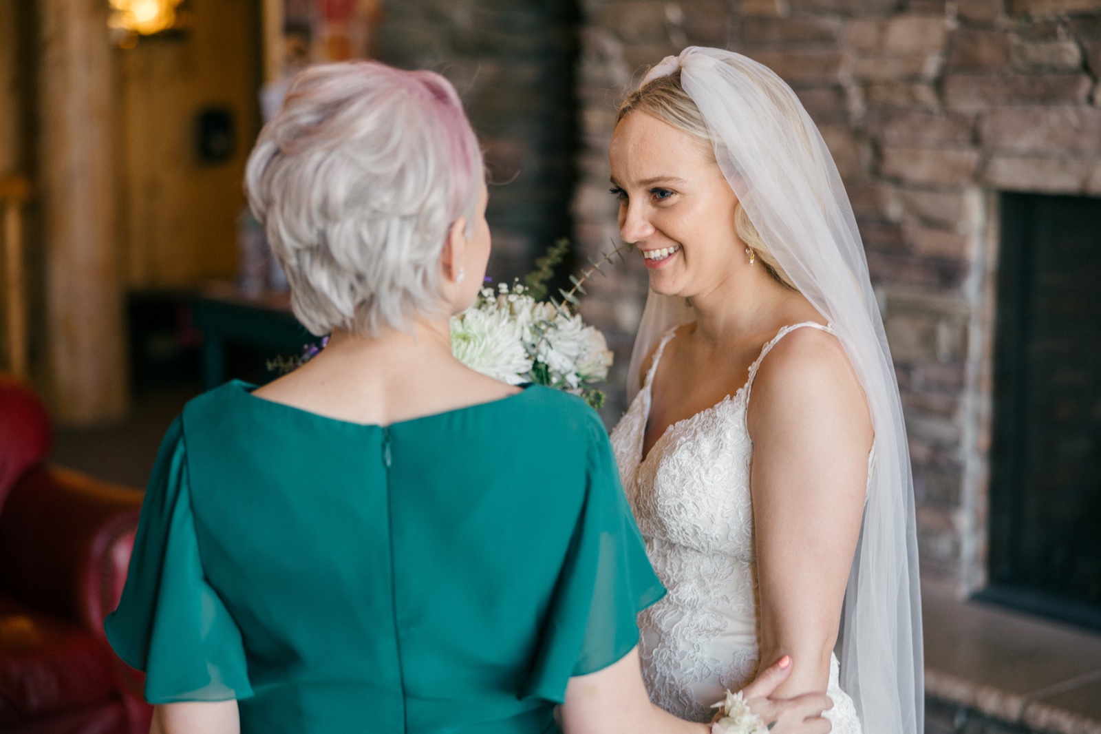 The bride in a lace gown and long veil smiles at a family member in a teal dress as they stand near the stone fireplace inside Whitefish Lodge — Tim Larsen Photography, Brainerd Lakes MN