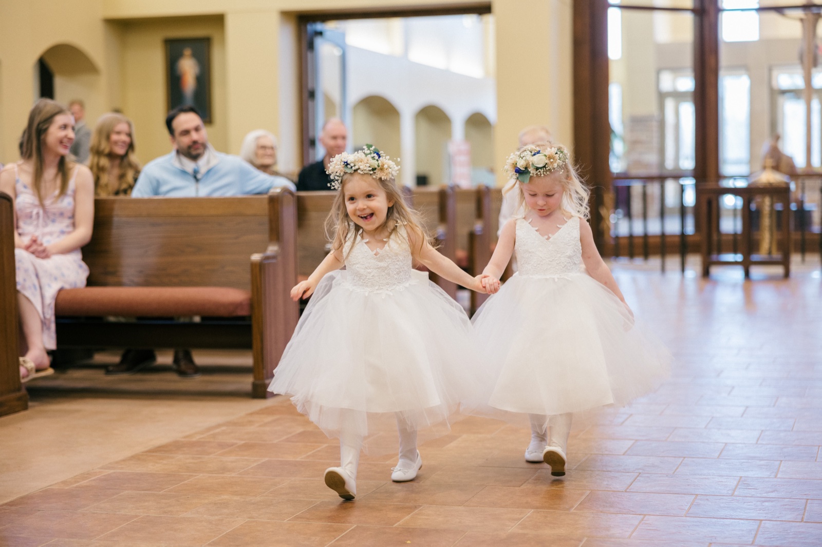 Two young flower girls in white tulle dresses and floral crowns hold hands as they walk down the church aisle, one beaming and the other looking down shyly — Tim Larsen Photography, Brainerd Lakes MN