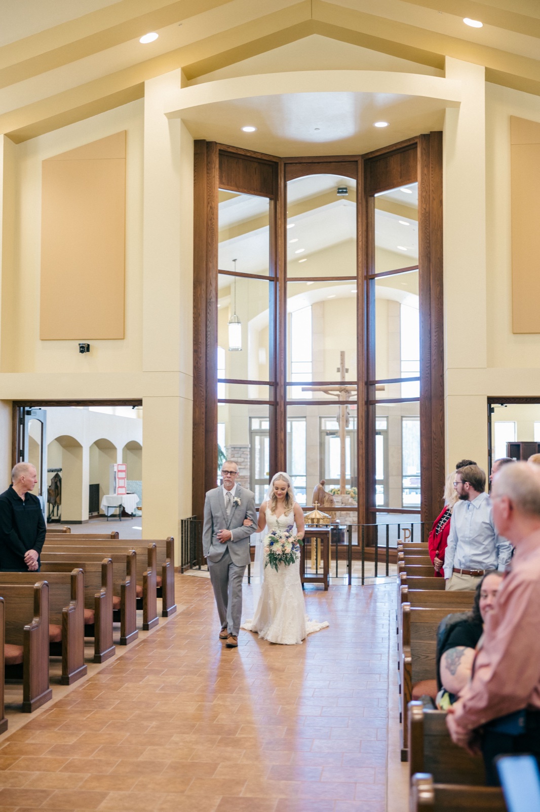 The bride walks down the aisle on her father's arm beneath the soaring wood-and-glass archway of the church sanctuary, with seated guests in the foreground pews — Tim Larsen Photography, Brainerd Lakes MN