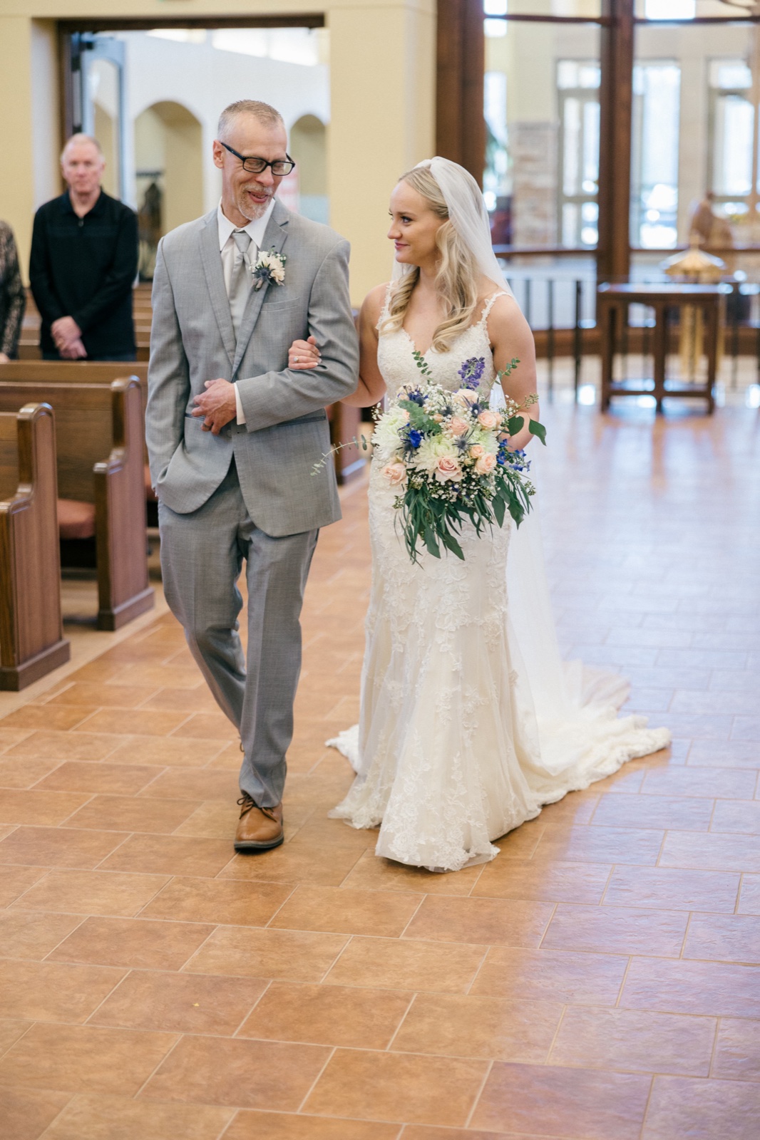 The bride walks down a tiled ceremony aisle on her father's arm, holding a large bouquet of pale roses and blue blooms, while a guest watches from a wooden pew — Tim Larsen Photography, Brainerd Lakes MN