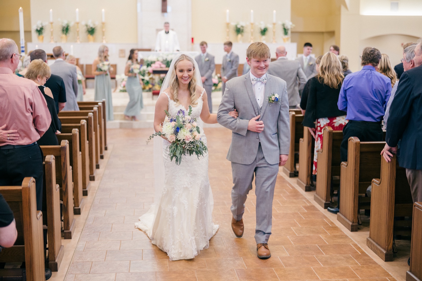The bride and groom walk down the church aisle together after their ceremony, the bride beaming and holding a pastel bouquet while the groom smiles beside her — Tim Larsen Photography, Brainerd Lakes MN