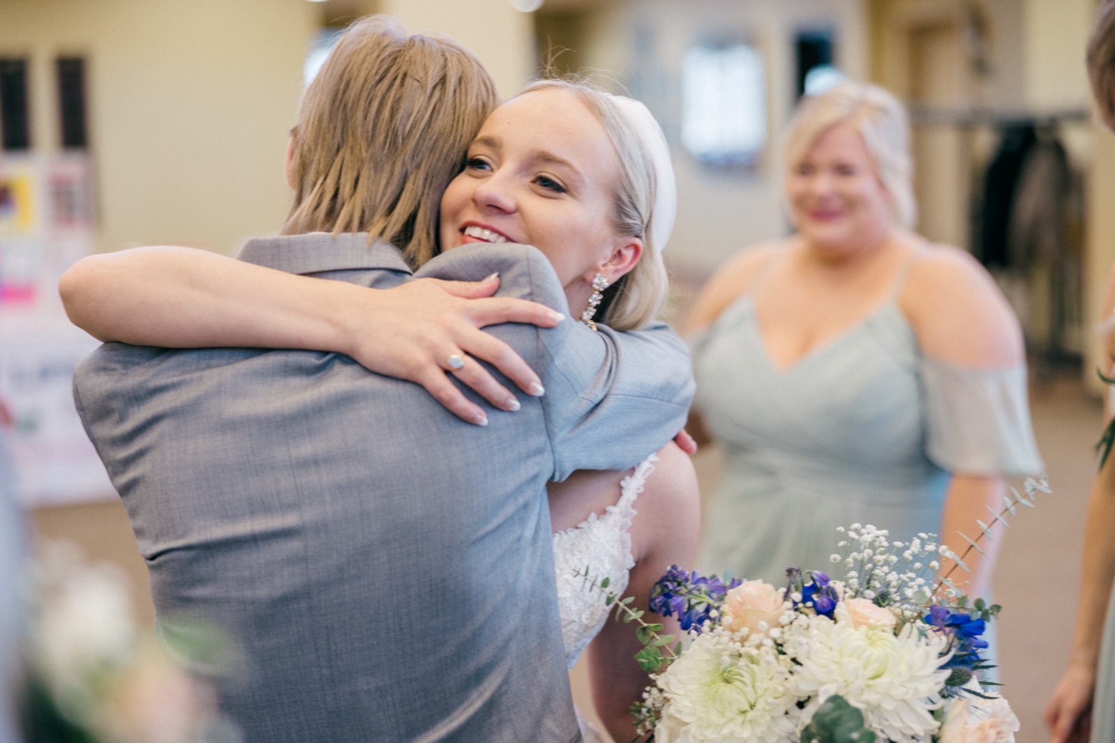 The bride beams over the shoulder of a guest she is hugging, while a bridesmaid in a sage dress smiles toward them holding a pastel bouquet — Tim Larsen Photography, Brainerd Lakes MN
