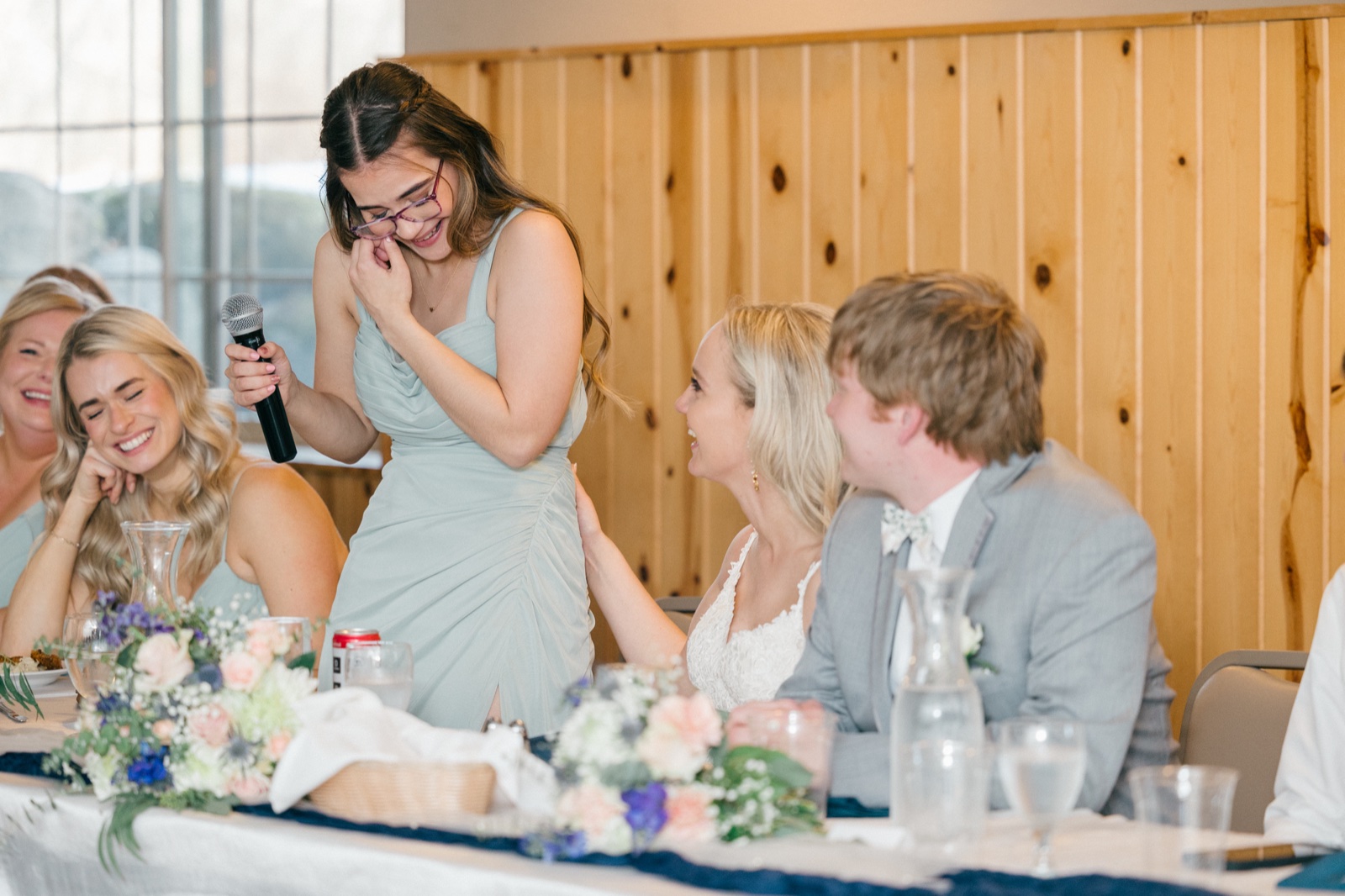 A bridesmaid in a sage dress holds a microphone and laughs into her hand mid-toast while the bride and groom laugh together at the head table — Tim Larsen Photography, Brainerd Lakes MN