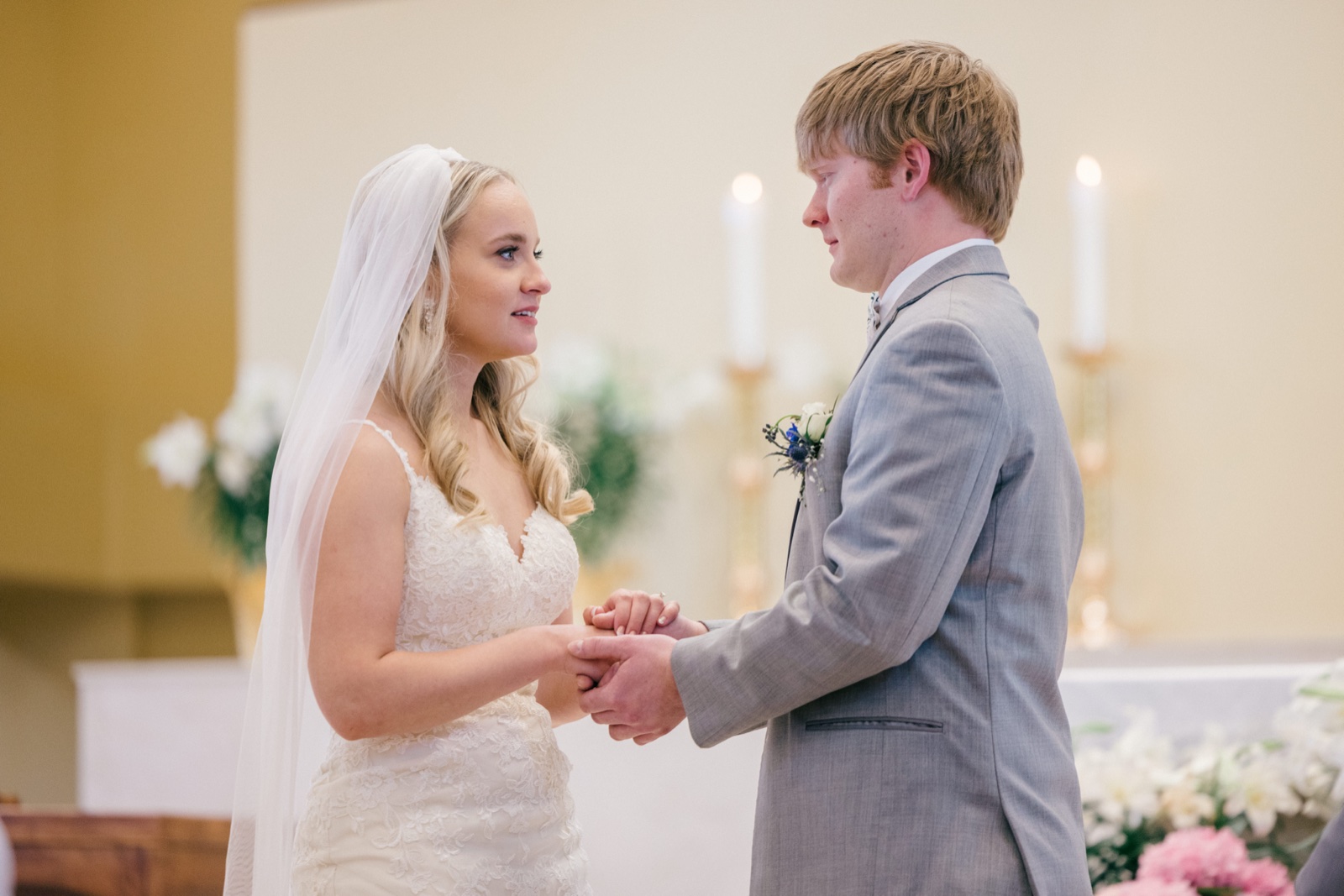 The bride and groom hold hands facing each other at the altar during their ceremony, with candles and floral arrangements softly visible behind them — Tim Larsen Photography, Brainerd Lakes MN