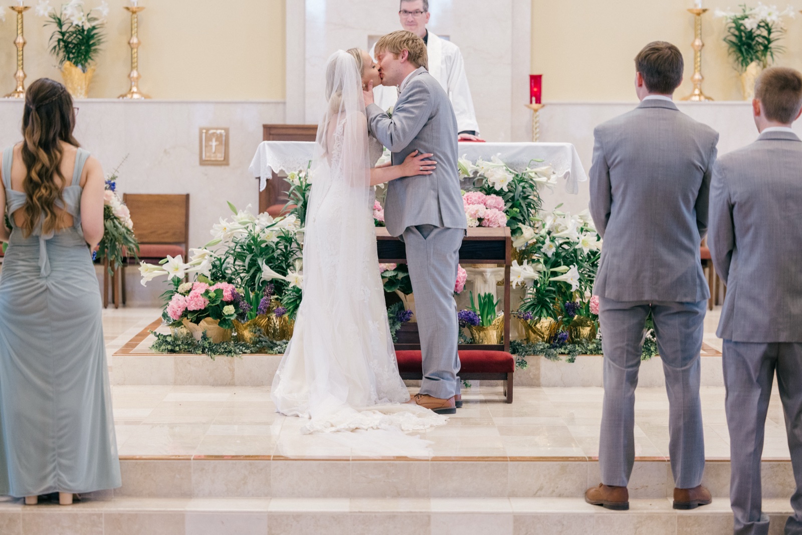 The bride and groom share their first kiss at the altar of the Catholic church surrounded by spring lilies and hydrangeas, framed by attendants on either side — Tim Larsen Photography, Brainerd Lakes MN