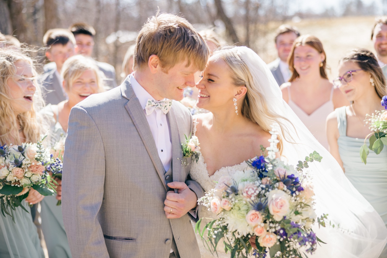 The bride and groom touch foreheads and smile at each other while their bridal party laughs around them in bright spring sunlight — Tim Larsen Photography, Brainerd Lakes MN