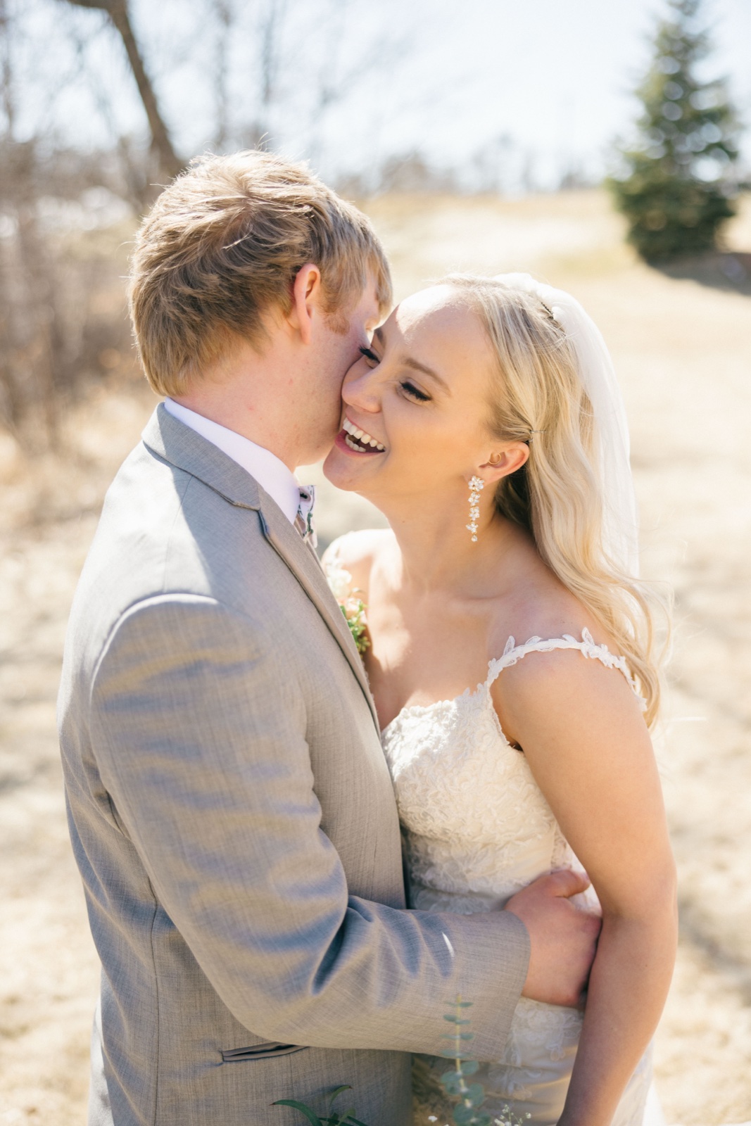 The groom presses his face to the bride's cheek as she laughs openly, her veil drifting behind her in an open field on a bright spring afternoon — Tim Larsen Photography, Brainerd Lakes MN