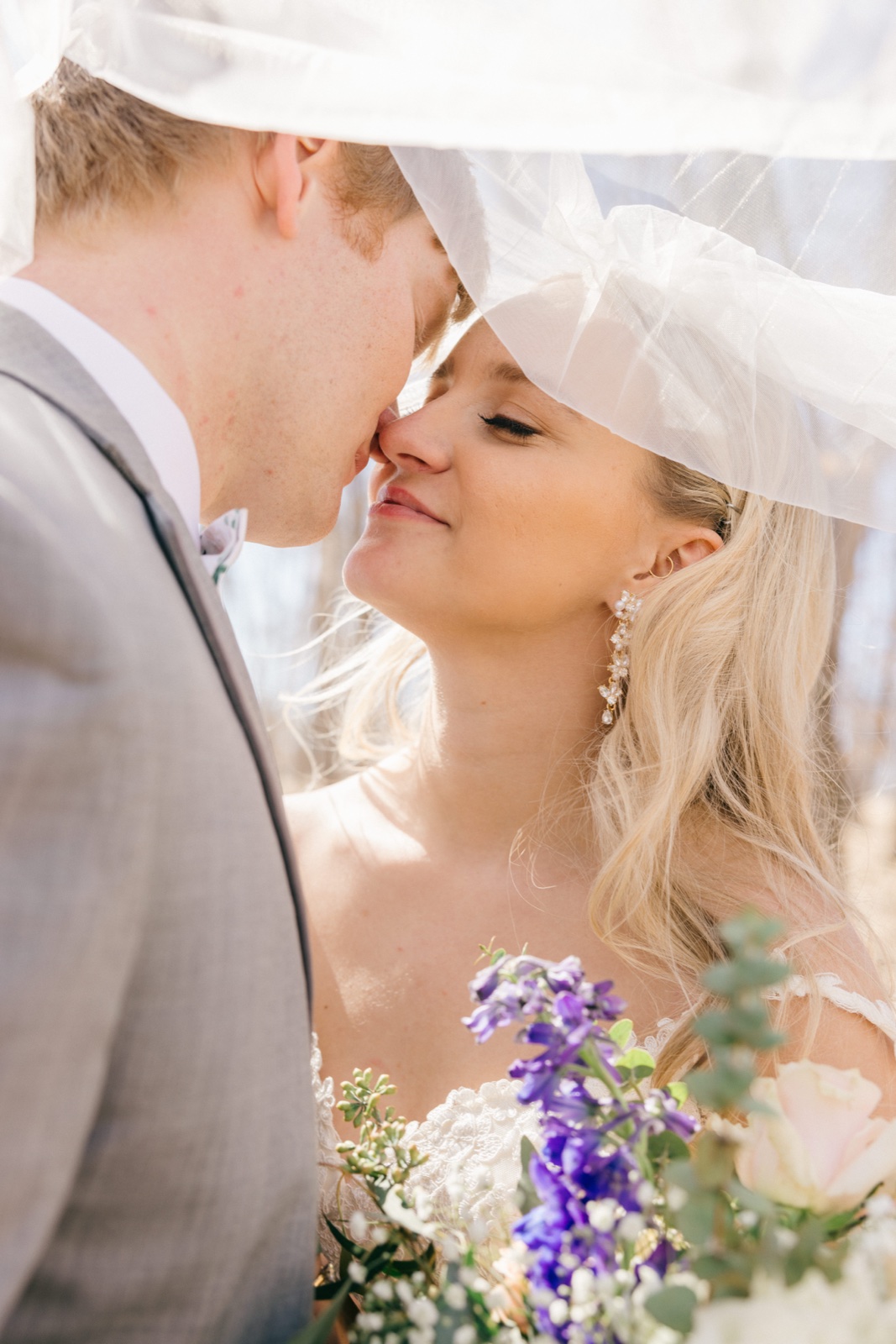 The bride and groom lean in close for a kiss beneath her veil, which drapes overhead lit translucent by the sun, her purple and white bouquet visible in the foreground — Tim Larsen Photography, Brainerd Lakes MN