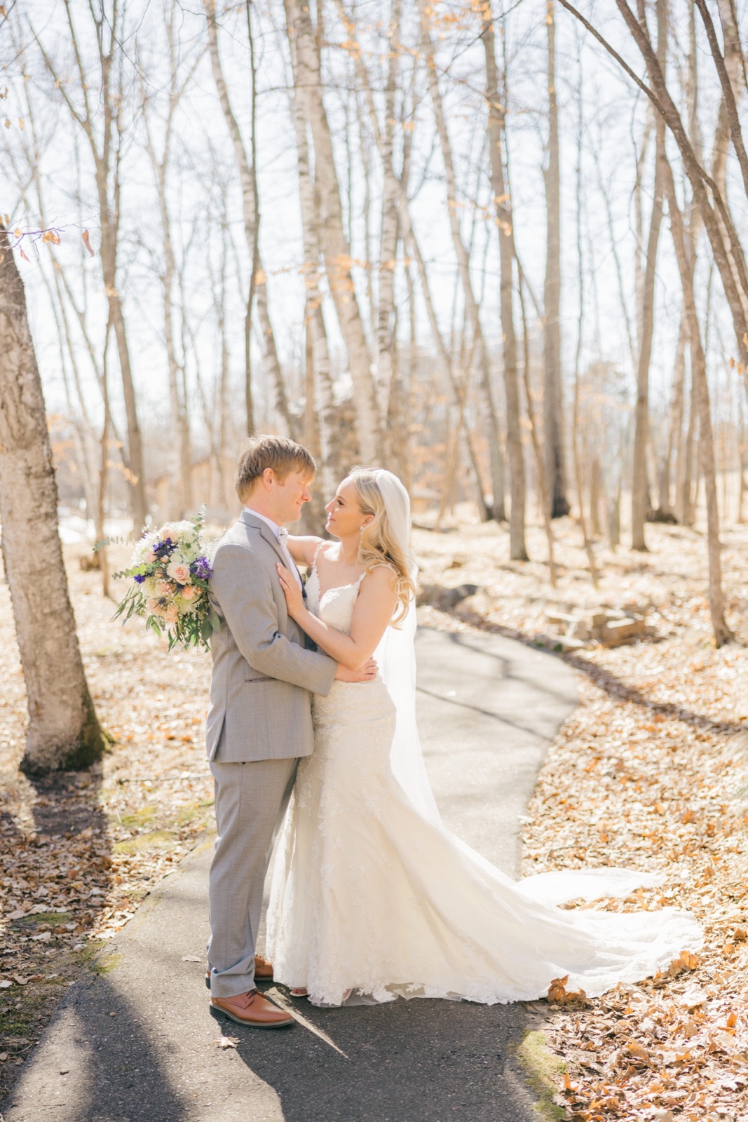 The bride and groom embrace on a sunlit leaf-covered path surrounded by tall bare birch trees, her gown's train trailing behind her — Tim Larsen Photography, Brainerd Lakes MN