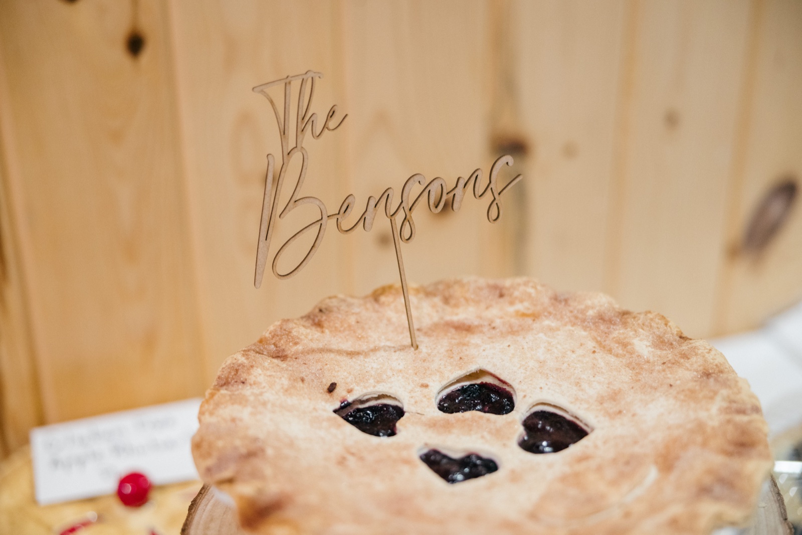 A laser-cut wooden cake topper reading 'The Bensons' stands in a blueberry pie with heart-shaped vents, against a pine-paneled wall — Tim Larsen Photography, Brainerd Lakes MN