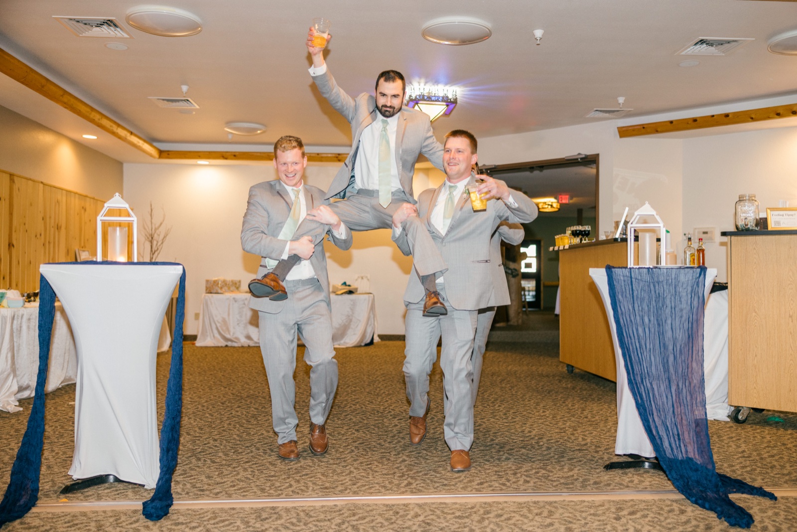 The groom is hoisted on the shoulders of two groomsmen, drink raised overhead, as they enter the reception room at Whitefish Lodge — Tim Larsen Photography, Brainerd Lakes MN