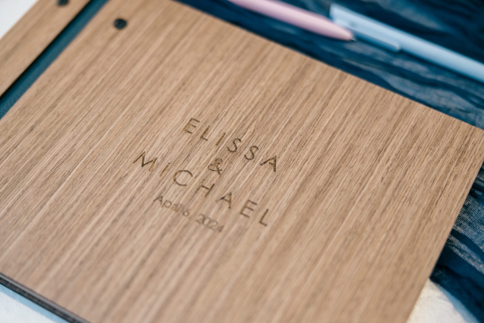 An engraved wood guest book reads with the couple's names beside softly out-of-focus pastel pens on dark fabric — Tim Larsen Photography, Brainerd Lakes MN
