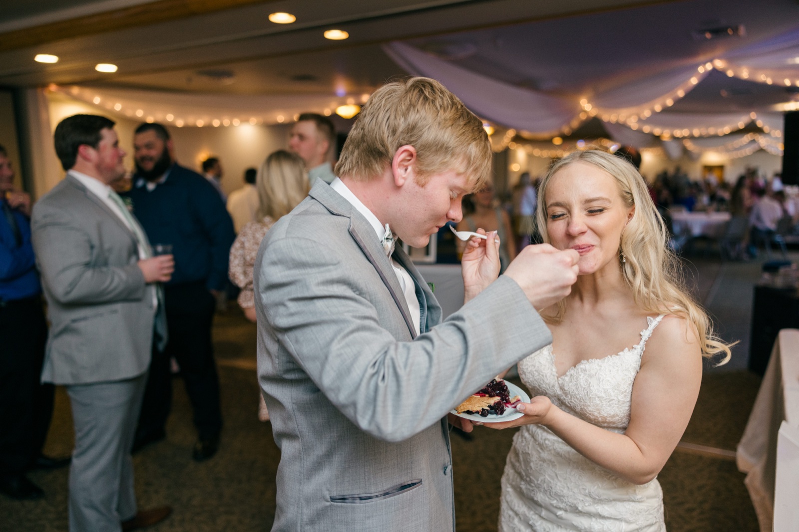 The bride scrunches her face playfully as she and the groom feed each other bites of pie at their reception, string lights glowing behind them — Tim Larsen Photography, Brainerd Lakes MN