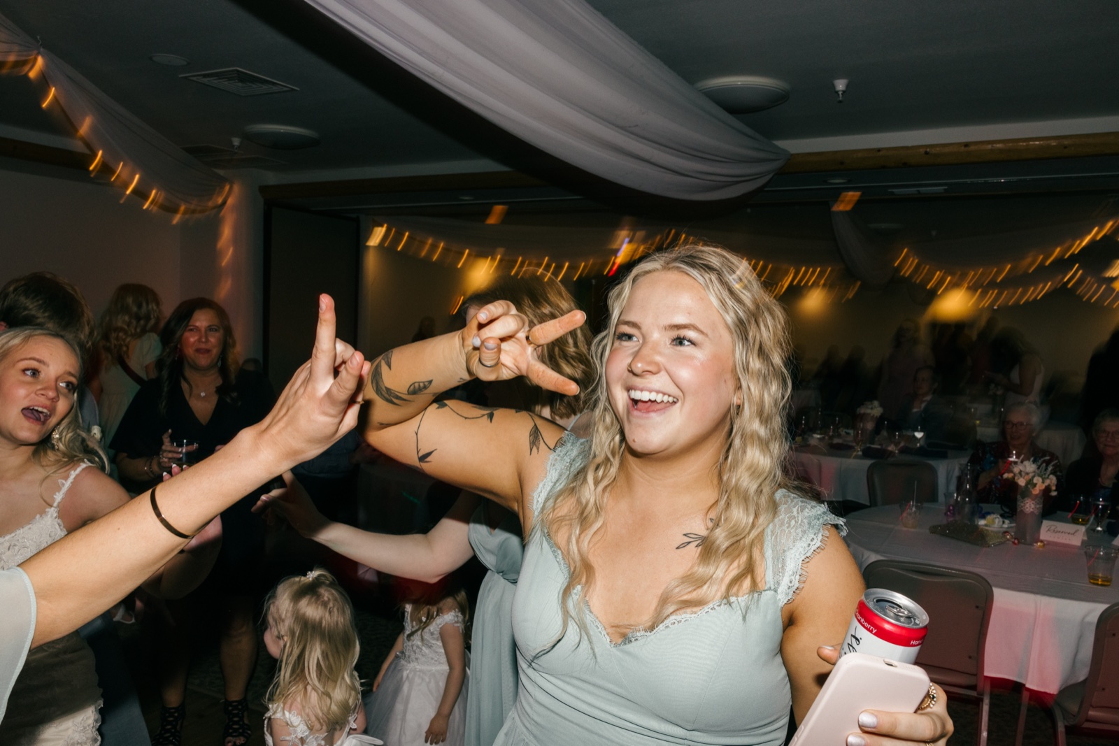 A bridesmaid in a sage dress laughs and throws up peace signs on the dance floor while holding a drink and phone, with guests and string lights behind her — Tim Larsen Photography, Brainerd Lakes MN