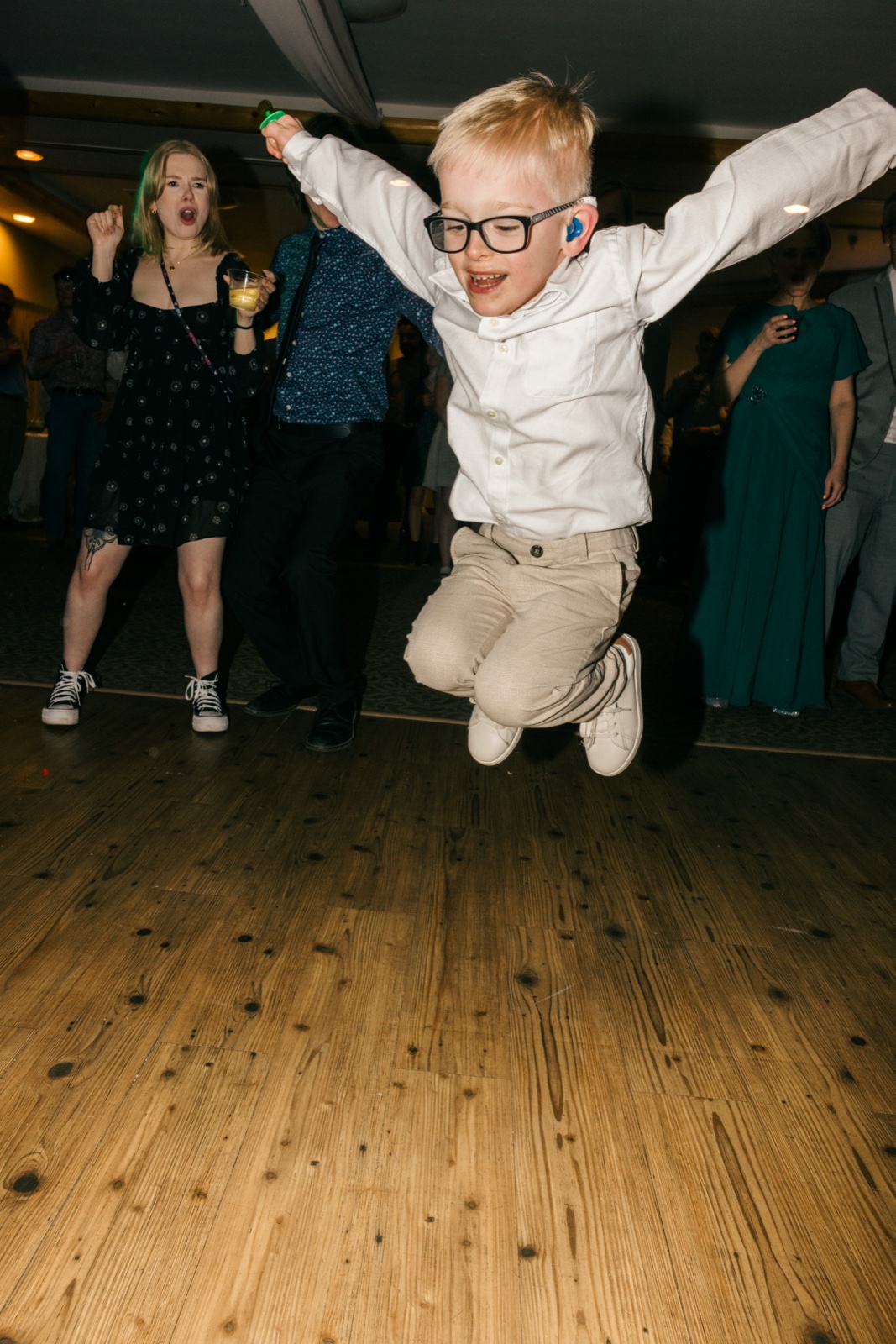 A young boy in glasses and a white button-down leaps mid-air on the dance floor with arms outstretched as a woman cheers behind him — Tim Larsen Photography, Brainerd Lakes MN