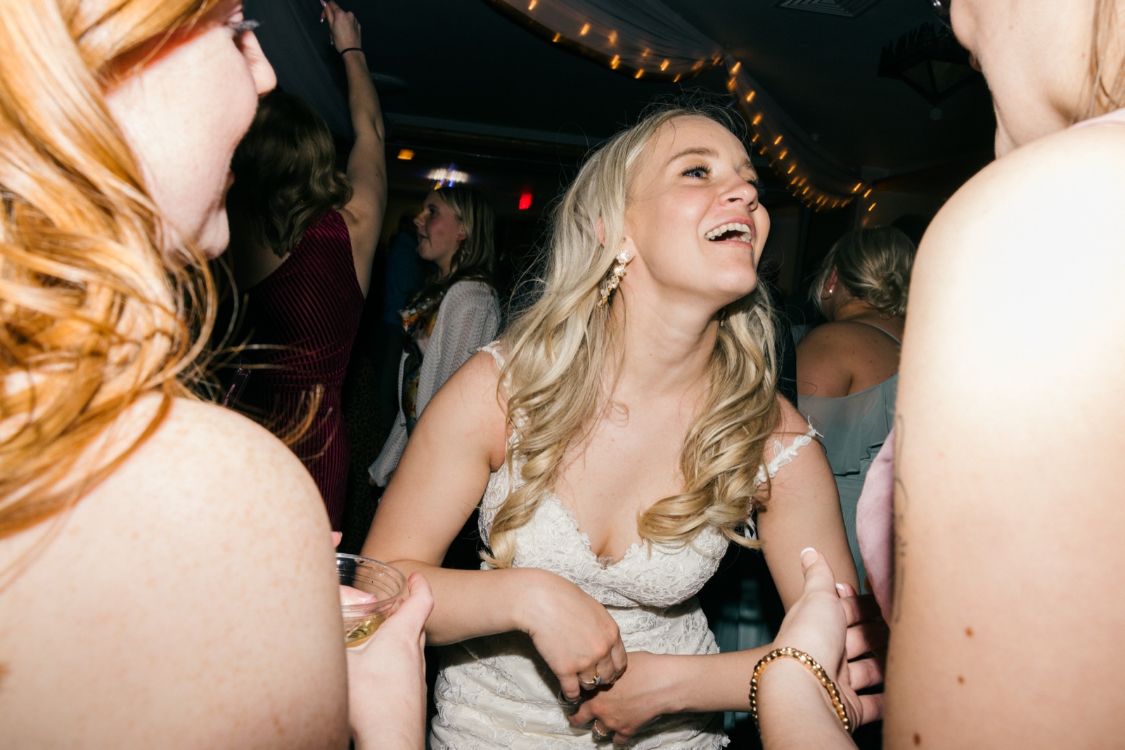 The bride laughs with her head tilted back while surrounded by friends on a dance floor lit by string lights overhead at Whitefish Lodge — Tim Larsen Photography, Brainerd Lakes MN