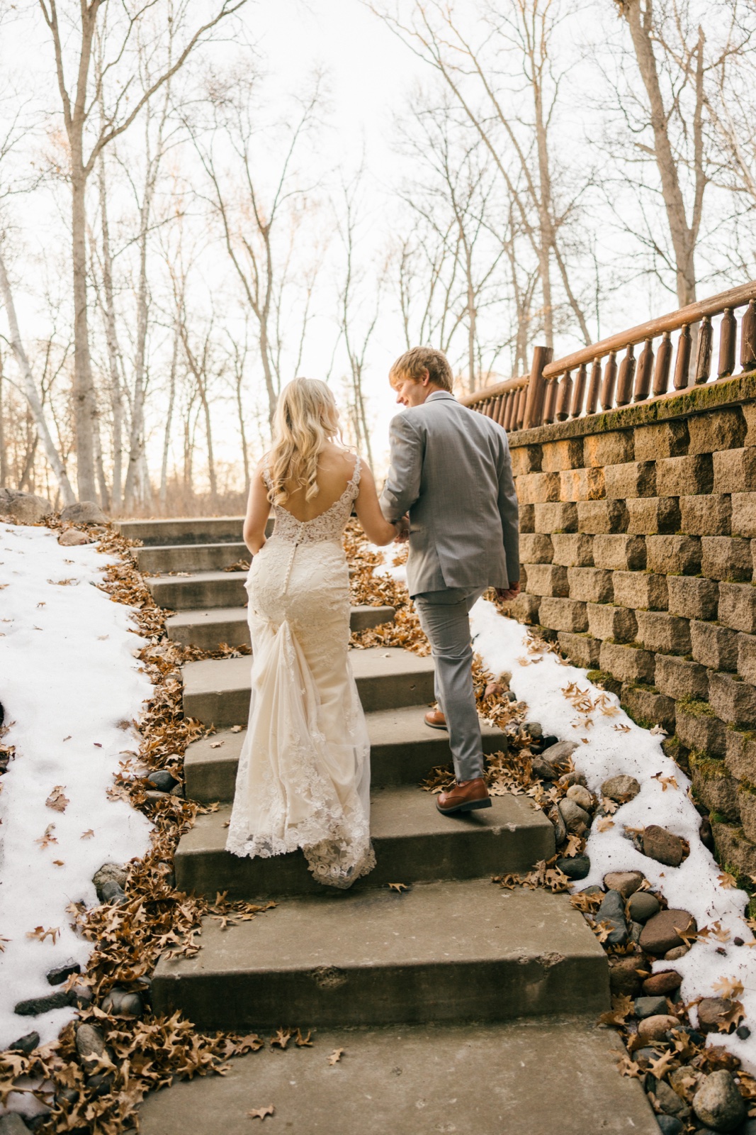 The bride and groom walk hand in hand up a stone staircase lined with fallen oak leaves and patches of snow, bare trees rising on either side — Tim Larsen Photography, Brainerd Lakes MN