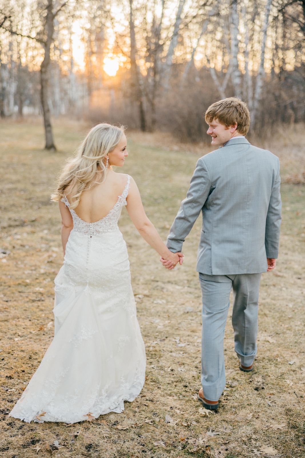 The bride and groom walk hand-in-hand away from the camera through a birch grove as the sun sets behind the trees, the bride glancing back toward her groom — Tim Larsen Photography, Brainerd Lakes MN