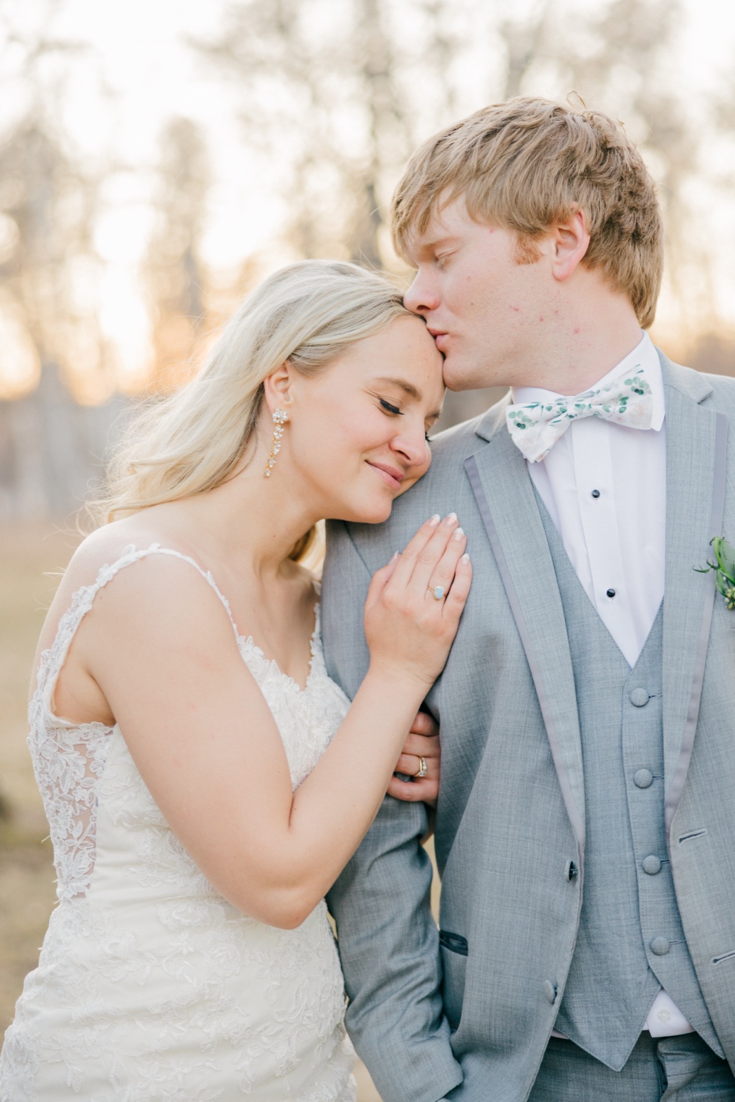 The groom kisses the bride's forehead as she rests against his shoulder with her hand on his chest, backlit by soft evening light through bare trees — Tim Larsen Photography, Brainerd Lakes MN