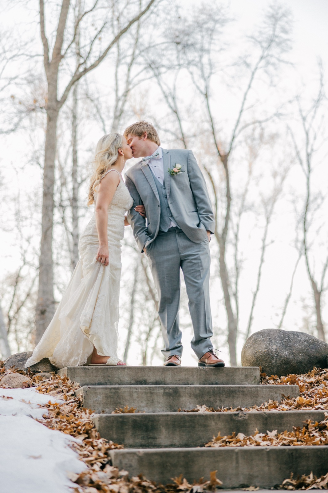 The couple shares a kiss at the top of a leaf-strewn stone staircase surrounded by bare trees and patches of snow at Whitefish Lodge — Tim Larsen Photography, Brainerd Lakes MN