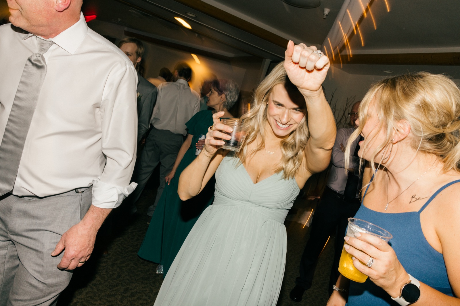 A bridesmaid in a sage green dress laughs and raises her fist in celebration on the dance floor while holding a drink, flanked by other guests — Tim Larsen Photography, Brainerd Lakes MN