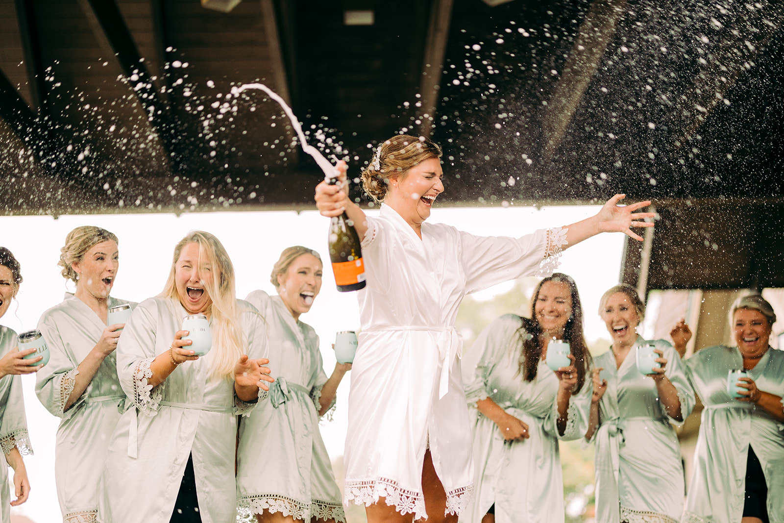 Bride popping champagne in a white robe while bridesmaids in matching sage green robes cheer — Tim Larsen Photography, Brainerd Lakes MN