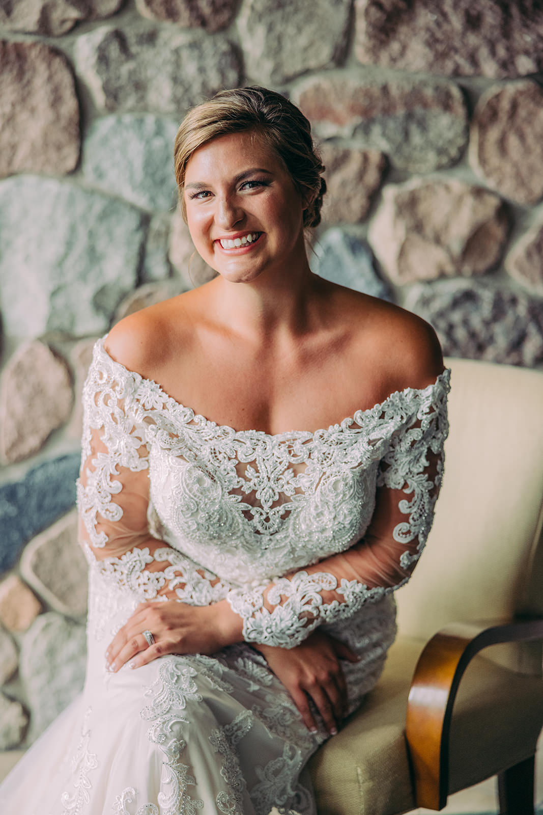 Bride in her off-shoulder long-sleeve lace gown seated against a stone wall — Tim Larsen Photography, Brainerd Lakes MN