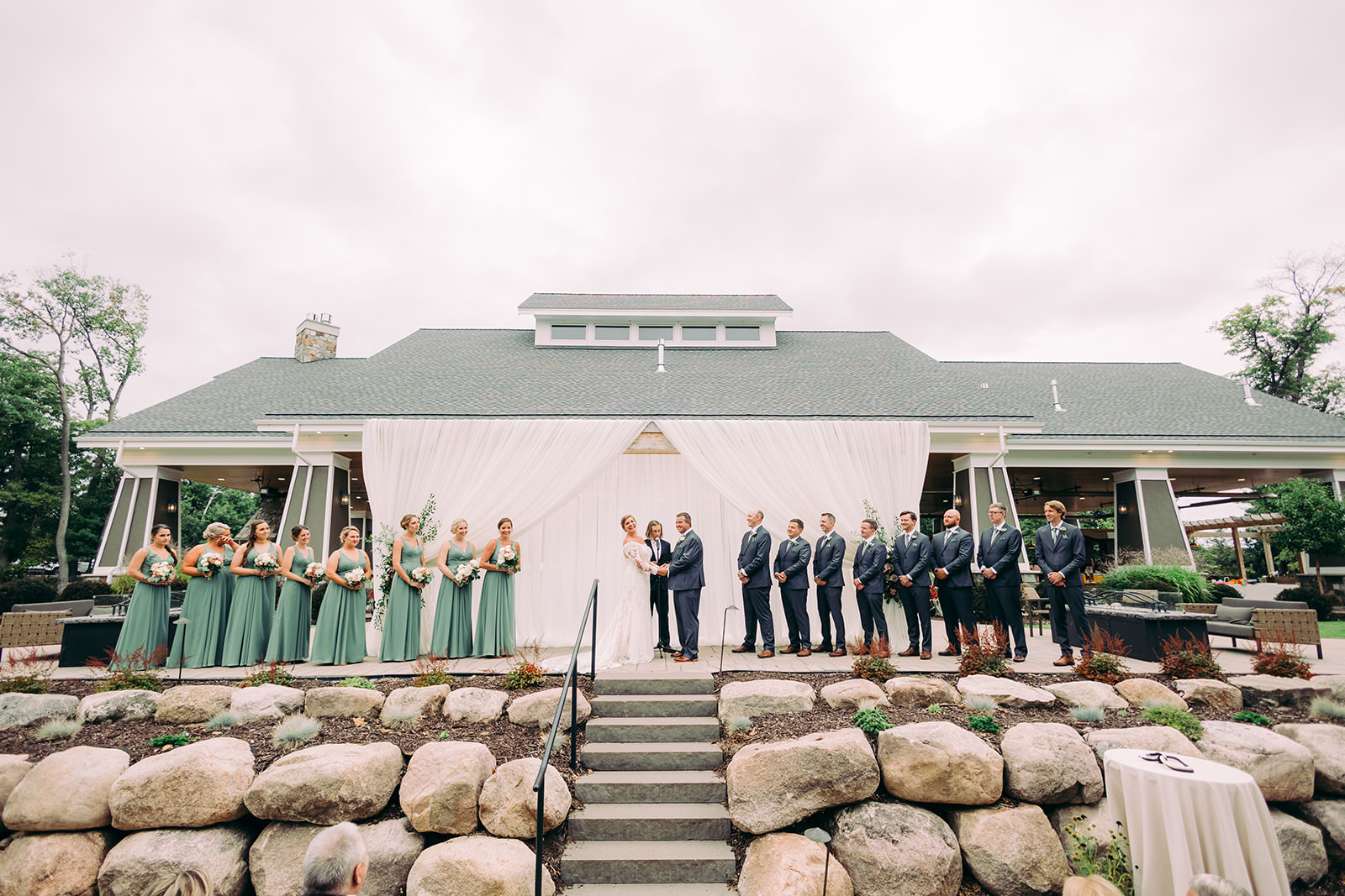 Wide shot of the outdoor ceremony at Madden's on Gull Lake — white-draped altar, bridal party along a stone retaining wall — Tim Larsen Photography, Brainerd Lakes MN