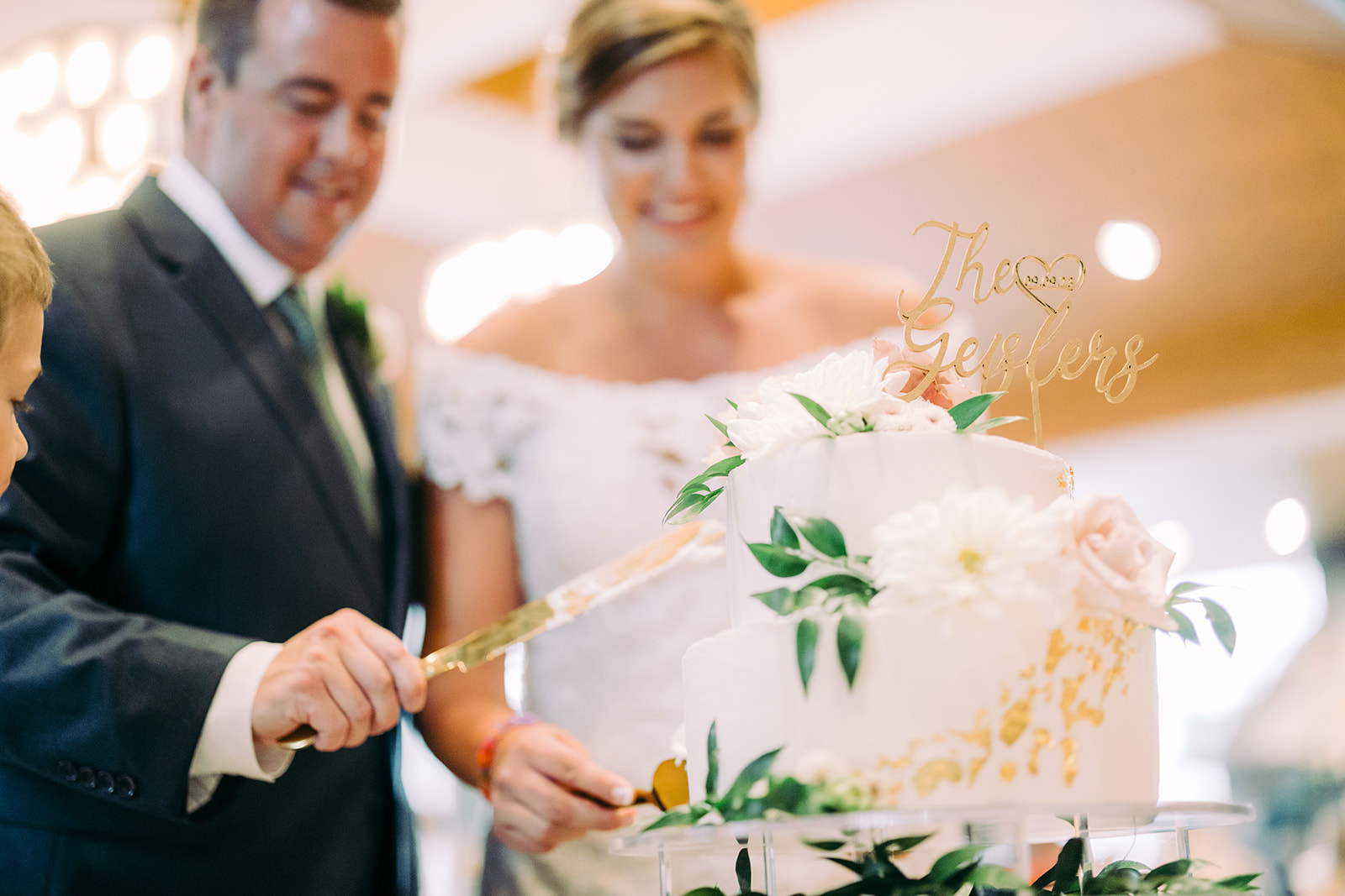 Cake cutting — custom gold "The Geislers" cake topper on a white tiered cake with cascading florals — Tim Larsen Photography, Brainerd Lakes MN