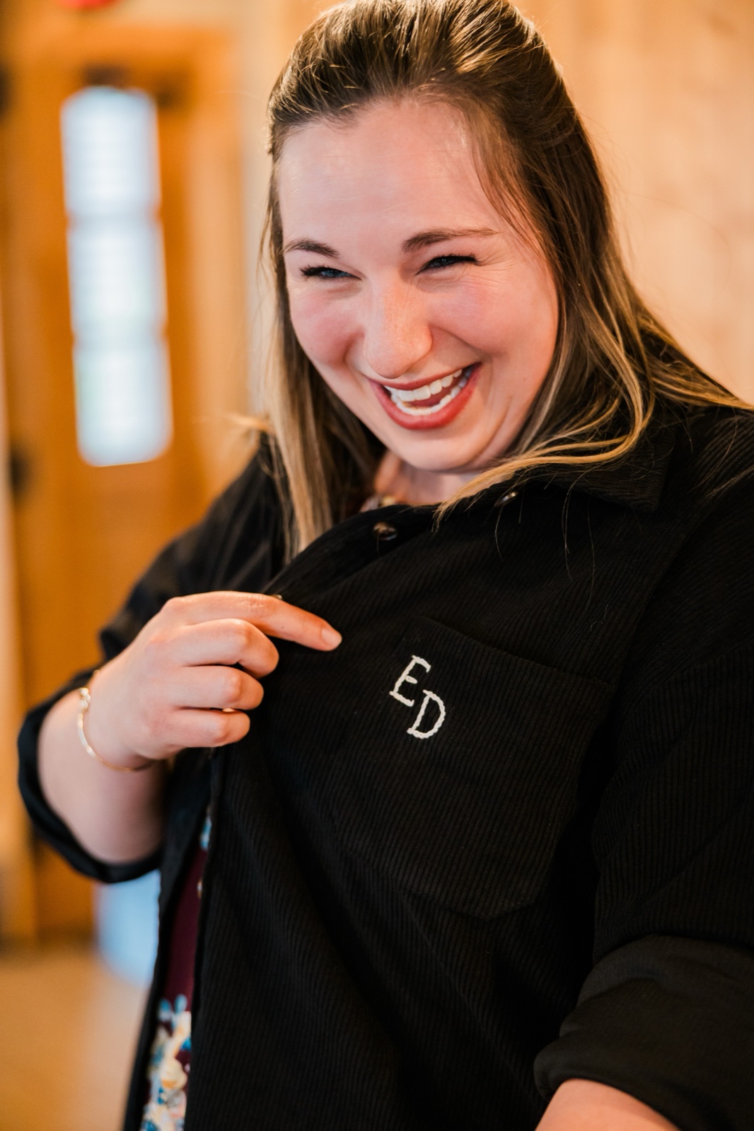 Bride pointing to the ED monogram on her black bridal jacket, mid-laugh in the Madden's cabin
