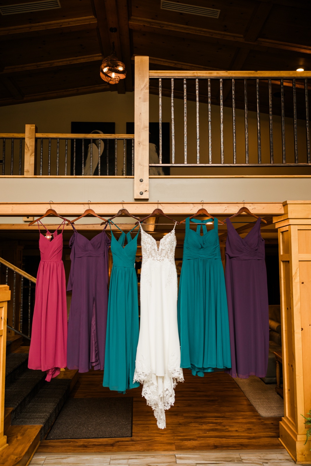 Bride's white lace gown hanging centered between six jewel-toned bridesmaid dresses on a cabin loft railing at Madden's on Gull Lake