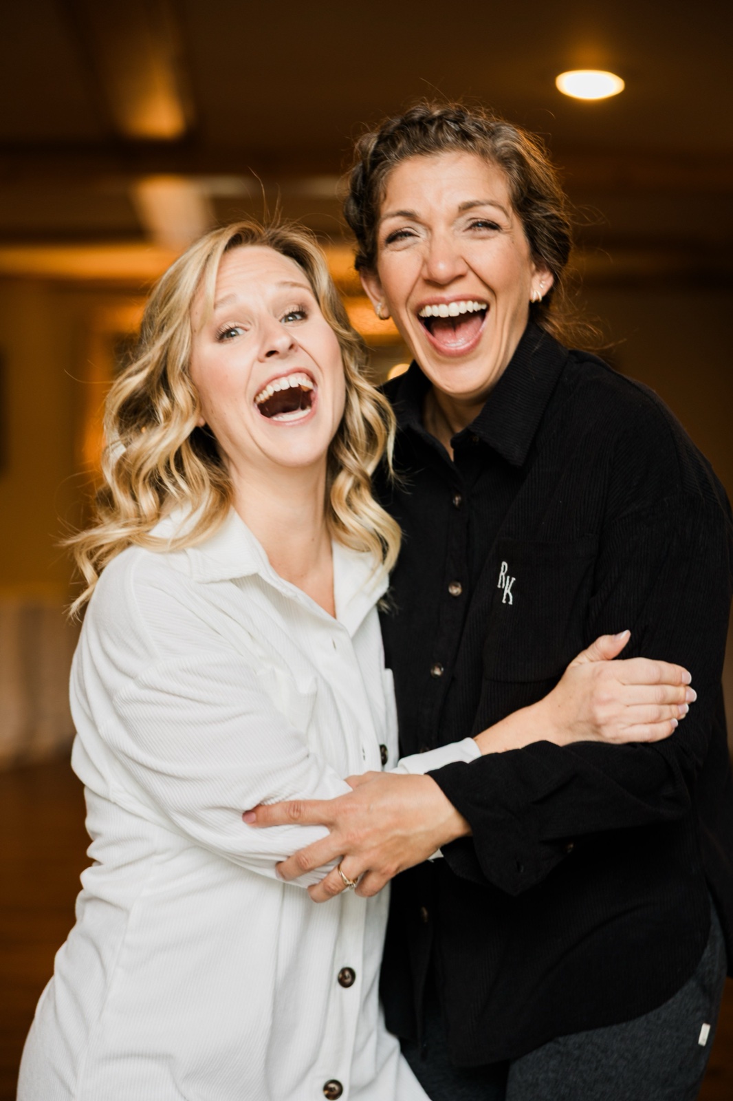 Bride and a close friend laughing together, arms wrapped around each other, in the warm reception room