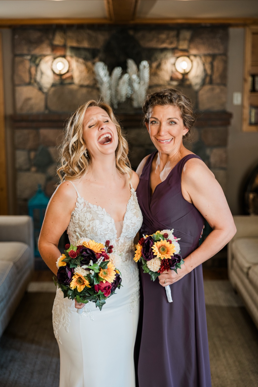 Bride laughing with a bridesmaid in deep purple in front of the stone fireplace at Madden's
