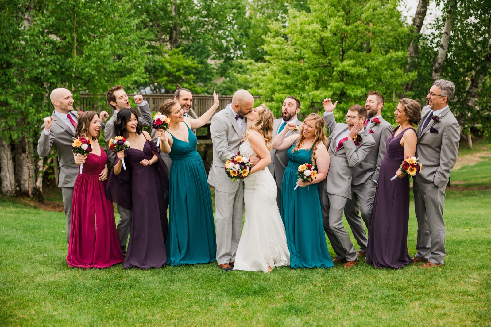 Bride and groom kissing surrounded by their cheering bridal party on a green lawn at Madden's