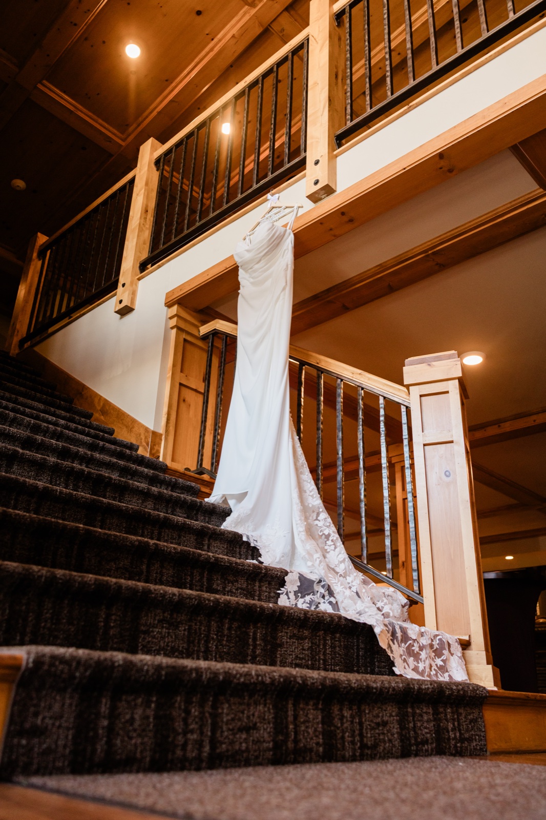White lace wedding dress hanging from a second-floor wooden railing with its long train cascading down a carpeted staircase inside the Madden's lodge — Tim Larsen Photography, Brainerd Lakes MN