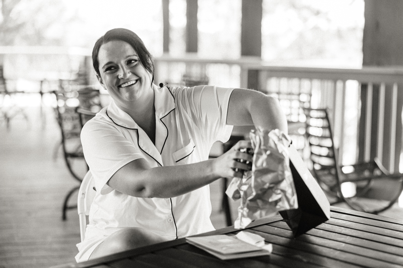 The bride laughing while unwrapping a gift bag on a covered porch lined with rocking chairs on the morning of her Madden's wedding — Tim Larsen Photography, Brainerd Lakes MN