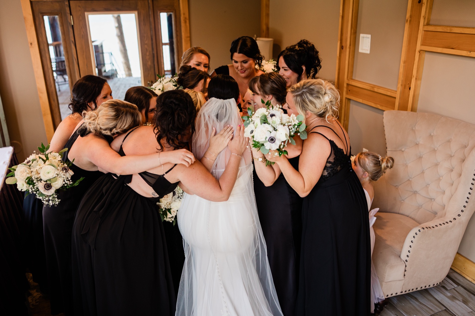 The bride surrounded in a close huddle by her bridesmaids, all holding white bouquets in the window-lit bridal suite at Madden's — Tim Larsen Photography, Brainerd Lakes MN