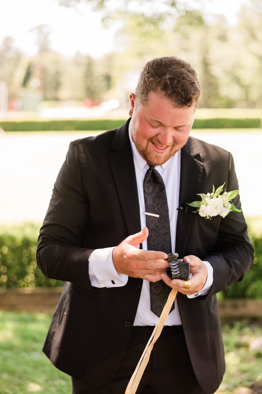 The groom in a black suit smiling at a watch he has just unwrapped, backlit on the resort lawn at Madden's — Tim Larsen Photography, Brainerd Lakes MN