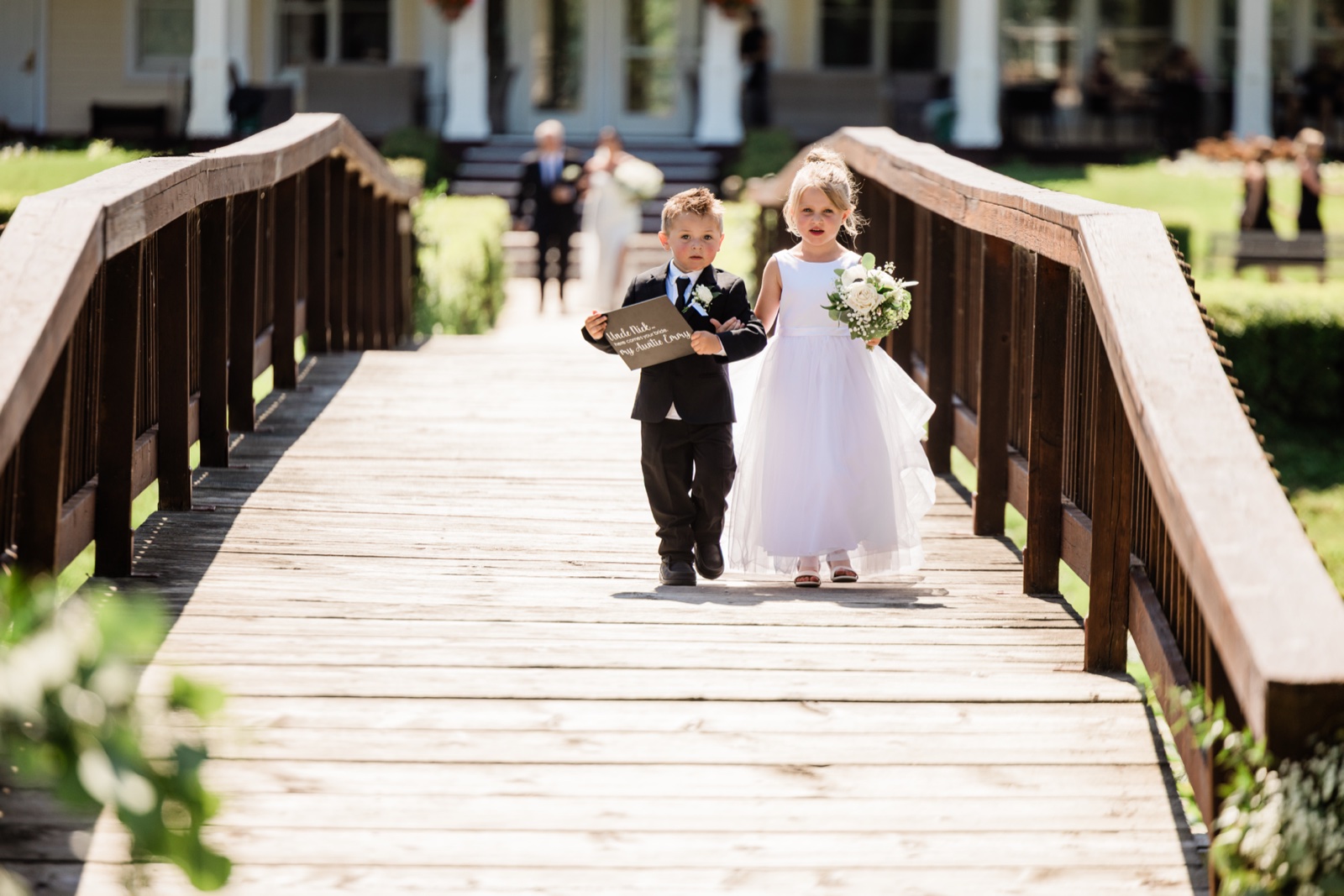 A young ring bearer and flower girl cross a wooden footbridge at Madden's arm-in-arm, carrying a sign and a small bouquet — Tim Larsen Photography, Brainerd Lakes MN