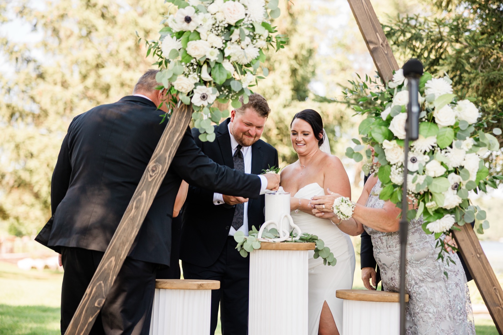The couple performs a unity sand ceremony at a pedestal beneath a wooden triangular arch draped with white florals on the Madden's lawn — Tim Larsen Photography, Brainerd Lakes MN