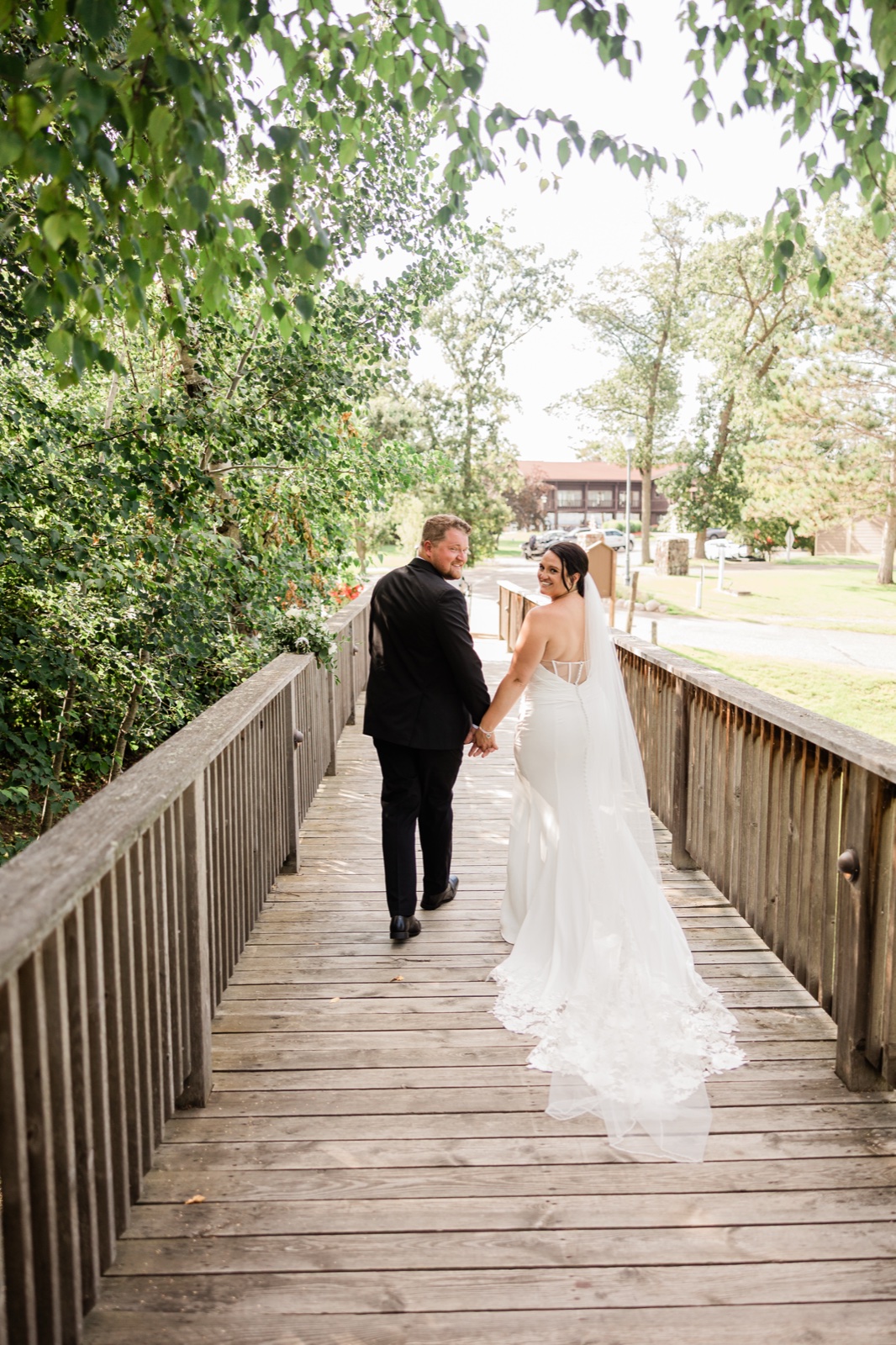 The bride glances back over her shoulder as she and the groom cross a wooden footbridge at Madden's, her veil trailing across the weathered planks — Tim Larsen Photography, Brainerd Lakes MN