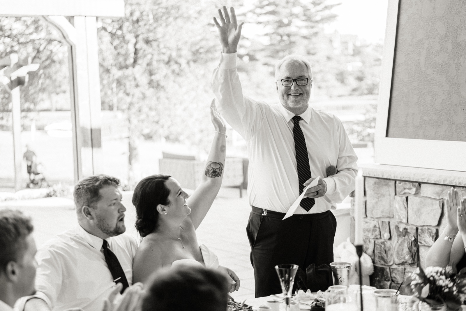 A man in glasses raises his hand mid-toast while the seated bride responds with a raised arm of her own — Tim Larsen Photography, Brainerd Lakes MN