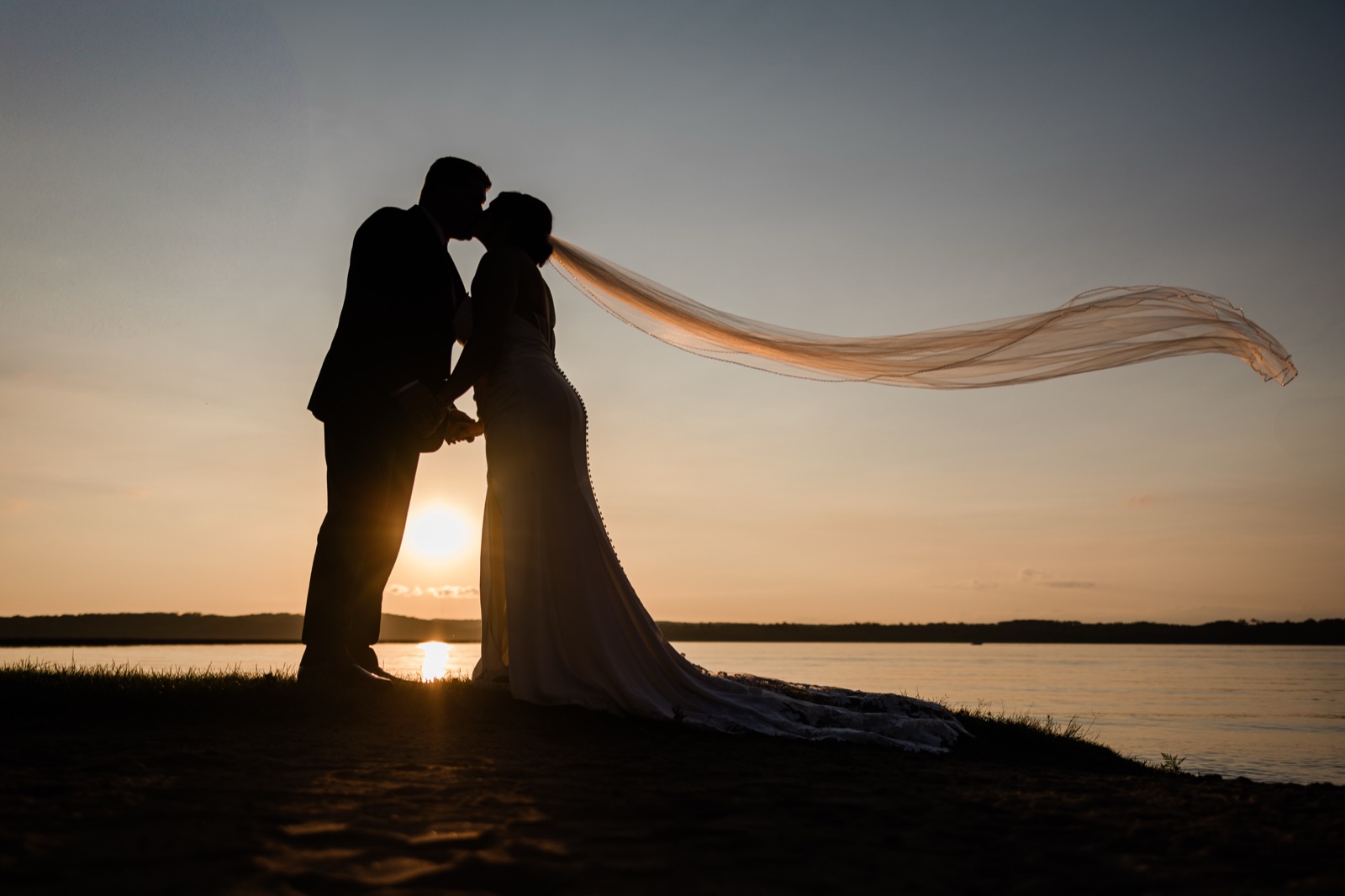 Silhouetted sunset kiss on the Gull Lake shore at Madden's, the bride's long veil lifting into a ribbon of light in the wind — Tim Larsen Photography, Brainerd Lakes MN