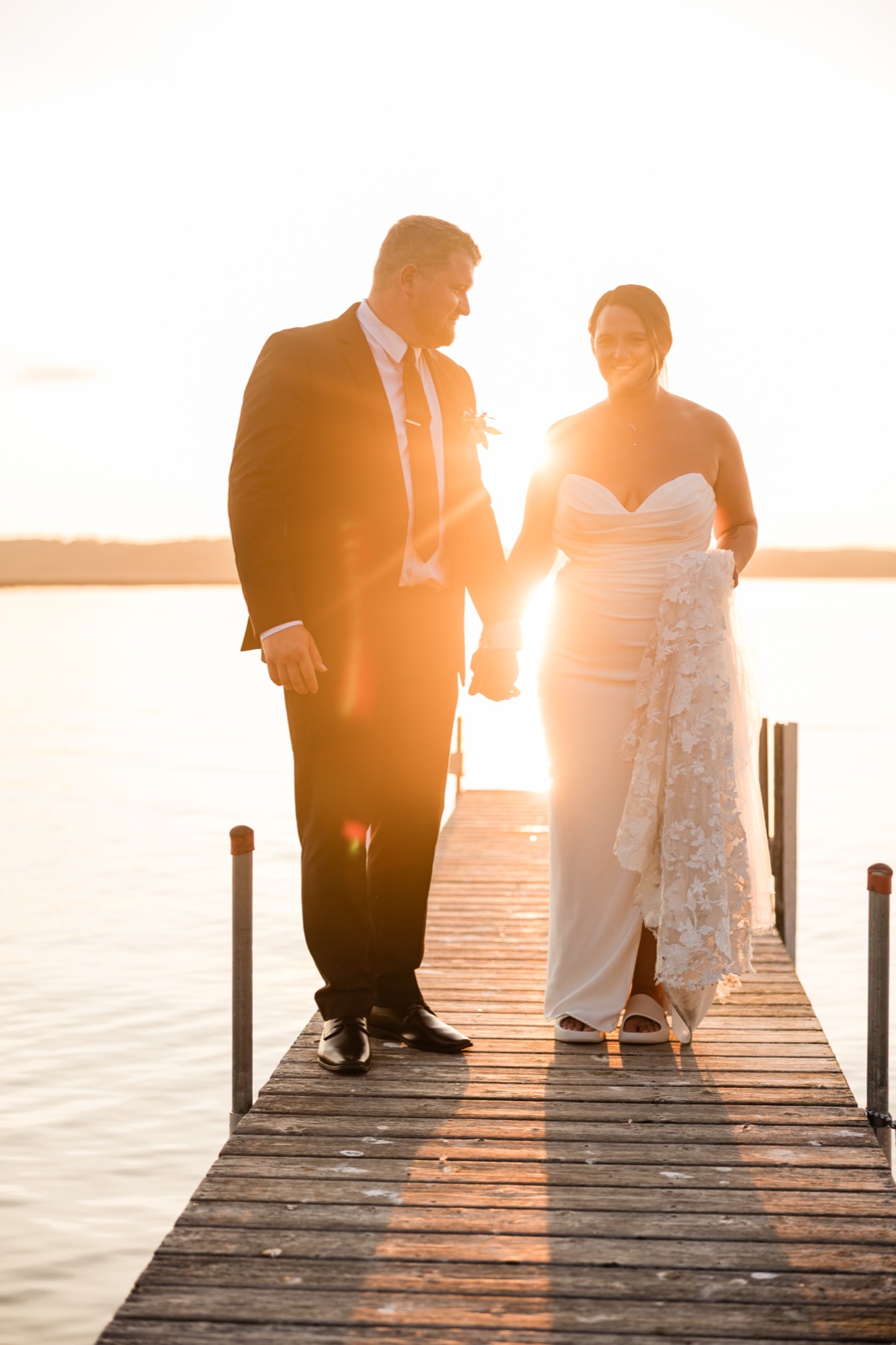 The bride and groom hold hands walking down a wooden dock on Gull Lake as sunset flares between them at Madden's — Tim Larsen Photography, Brainerd Lakes MN
