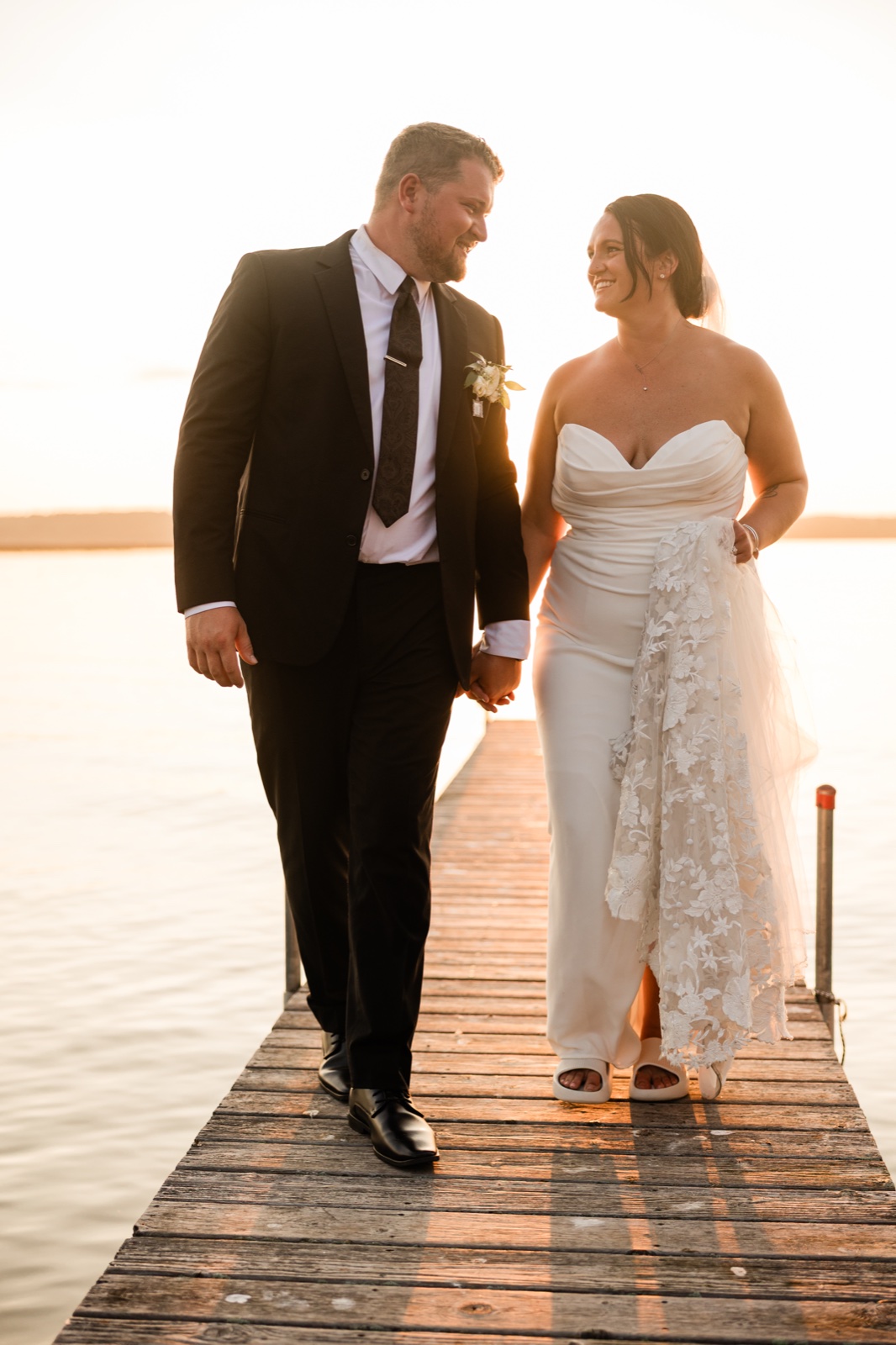 The couple walks toward the camera down a weathered wooden dock backlit by golden sunset light over Gull Lake — Tim Larsen Photography, Brainerd Lakes MN