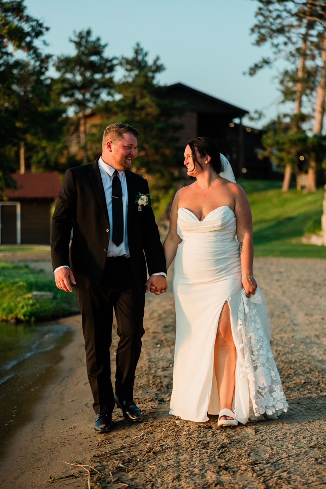 The bride and groom walk hand-in-hand along a sandy Gull Lake shoreline at golden hour, the Madden's lodge visible through the pines — Tim Larsen Photography, Brainerd Lakes MN