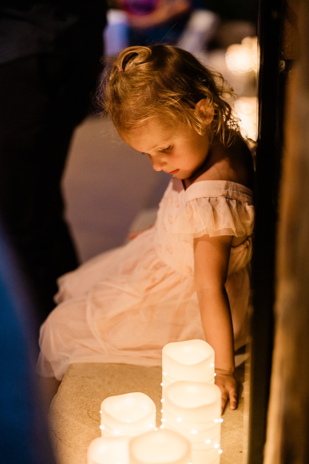 A young flower girl leans toward a cluster of glowing pillar candles, warmly lit in the dark Madden's reception space — Tim Larsen Photography, Brainerd Lakes MN
