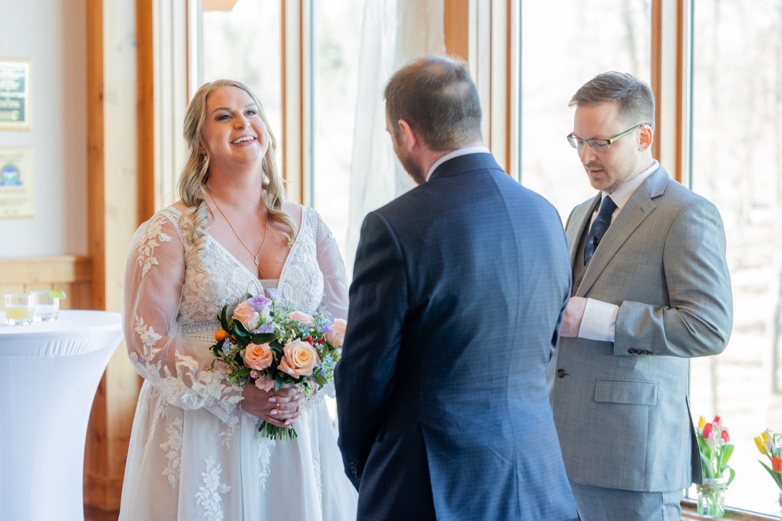 The bride smiles at the groom while holding her bouquet during the indoor ceremony at Cragun's Resort, spring woods filling the windows behind them — Tim Larsen Photography, Brainerd Lakes MN