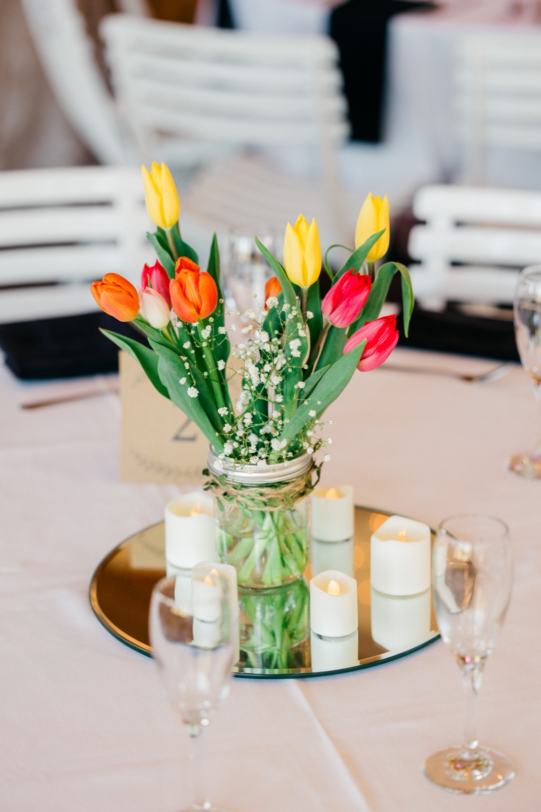 A mason jar centerpiece of orange, yellow, and pink tulips with baby's breath ringed by candlelight on a mirrored base at a Cragun's reception table — Tim Larsen Photography, Brainerd Lakes MN