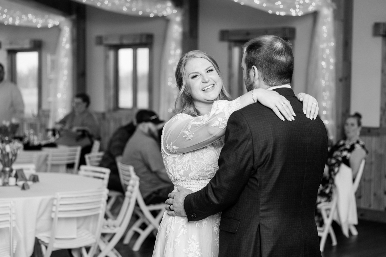 The bride smiles broadly over the groom's shoulder during their first dance, with string lights softly out of focus behind them — Tim Larsen Photography, Brainerd Lakes MN